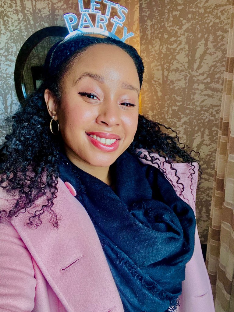 Smiling woman in a pink wool coat and scarf, facing morning sunlight and wearing a New Year headband in a cozy winter setting.